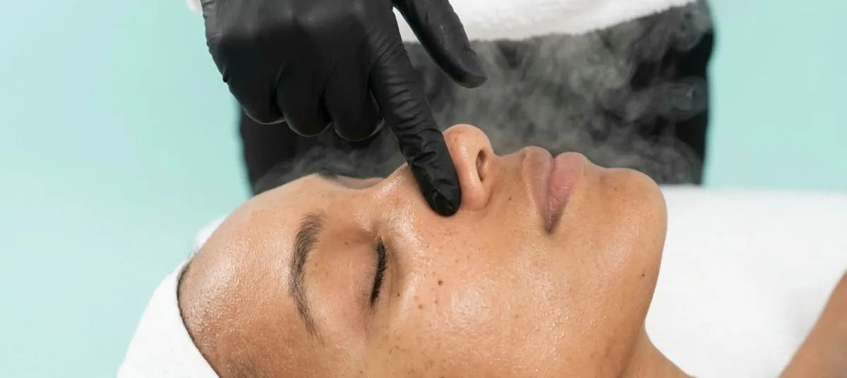 Esthetician at KiWest Spa Belleville performing the steam and extraction step of a deep cleansing facial on a client with freckled, melanin-rich skin in Barbados