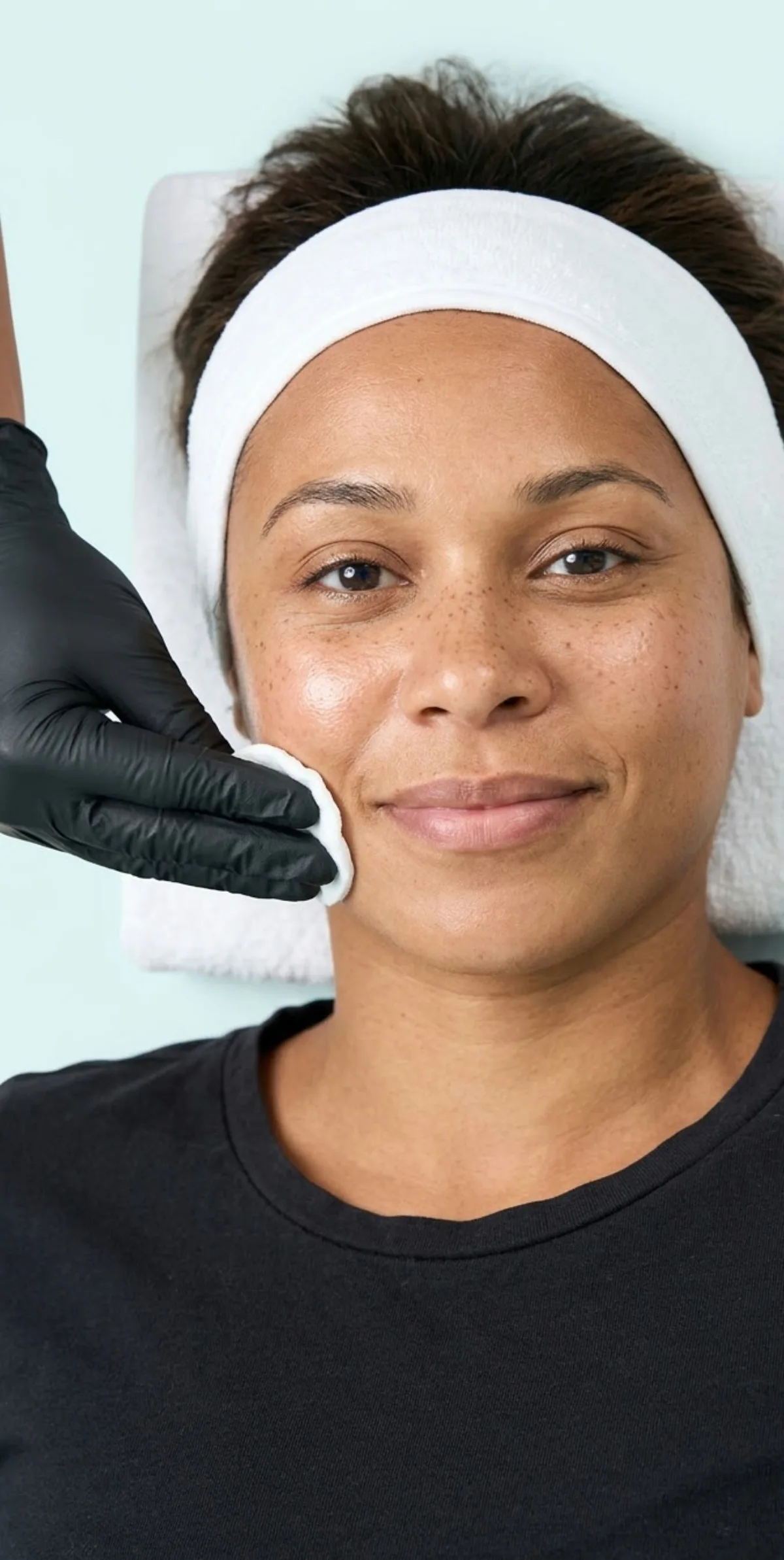 Esthetician at KiWest Spa Belleville pressing a toner-soaked cotton round to the cheek of a smiling Caribbean client with freckles during a 30-minute mini facial in Barbados