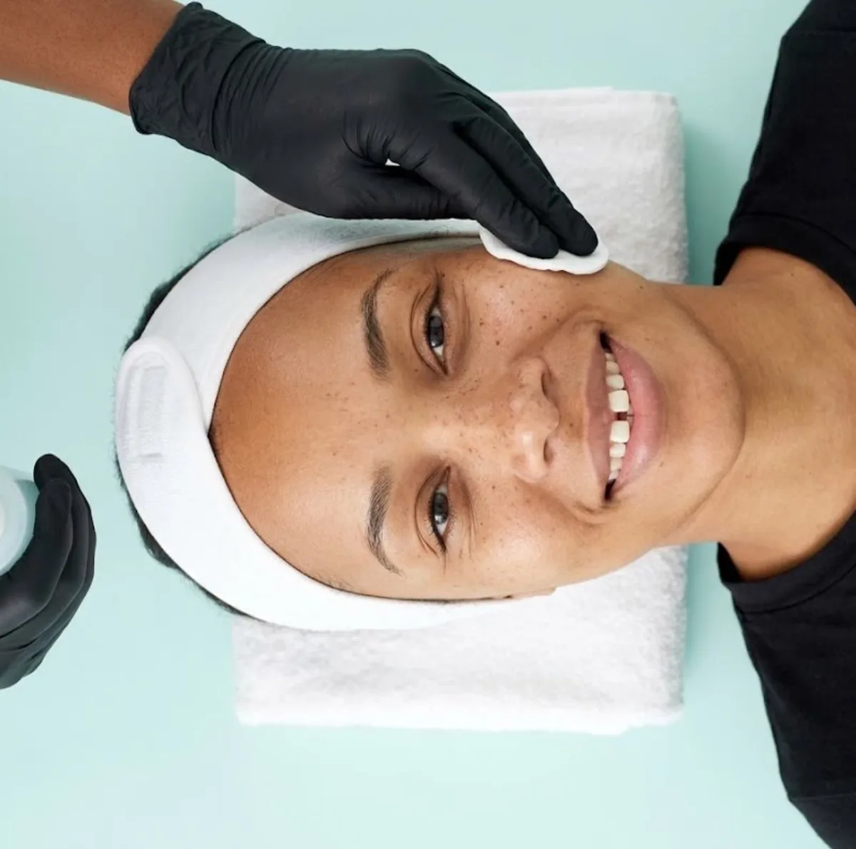 KiWest Spa esthetician applying a toner-soaked cotton round to the cheek of a smiling Caribbean client with freckles and a natural tooth gap during a 30-minute mini facial in Belleville, Barbados